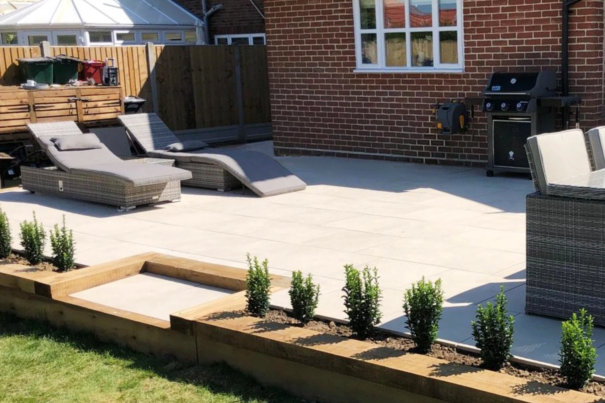 Contemporary porcelain paving patio installed in a rear garden with lounge chairs, outdoor dining furniture and raised timber borders.